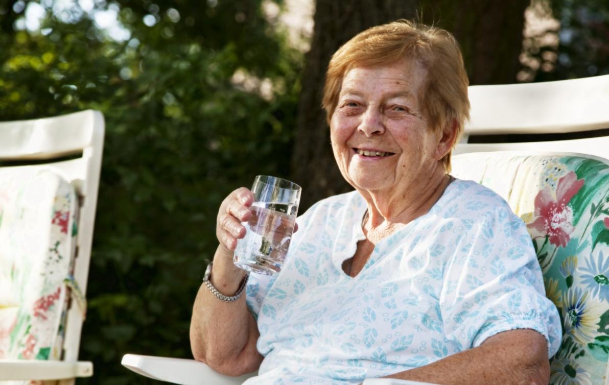 Athena is an elderly woman smiling at the camera whilst holding a glass of water