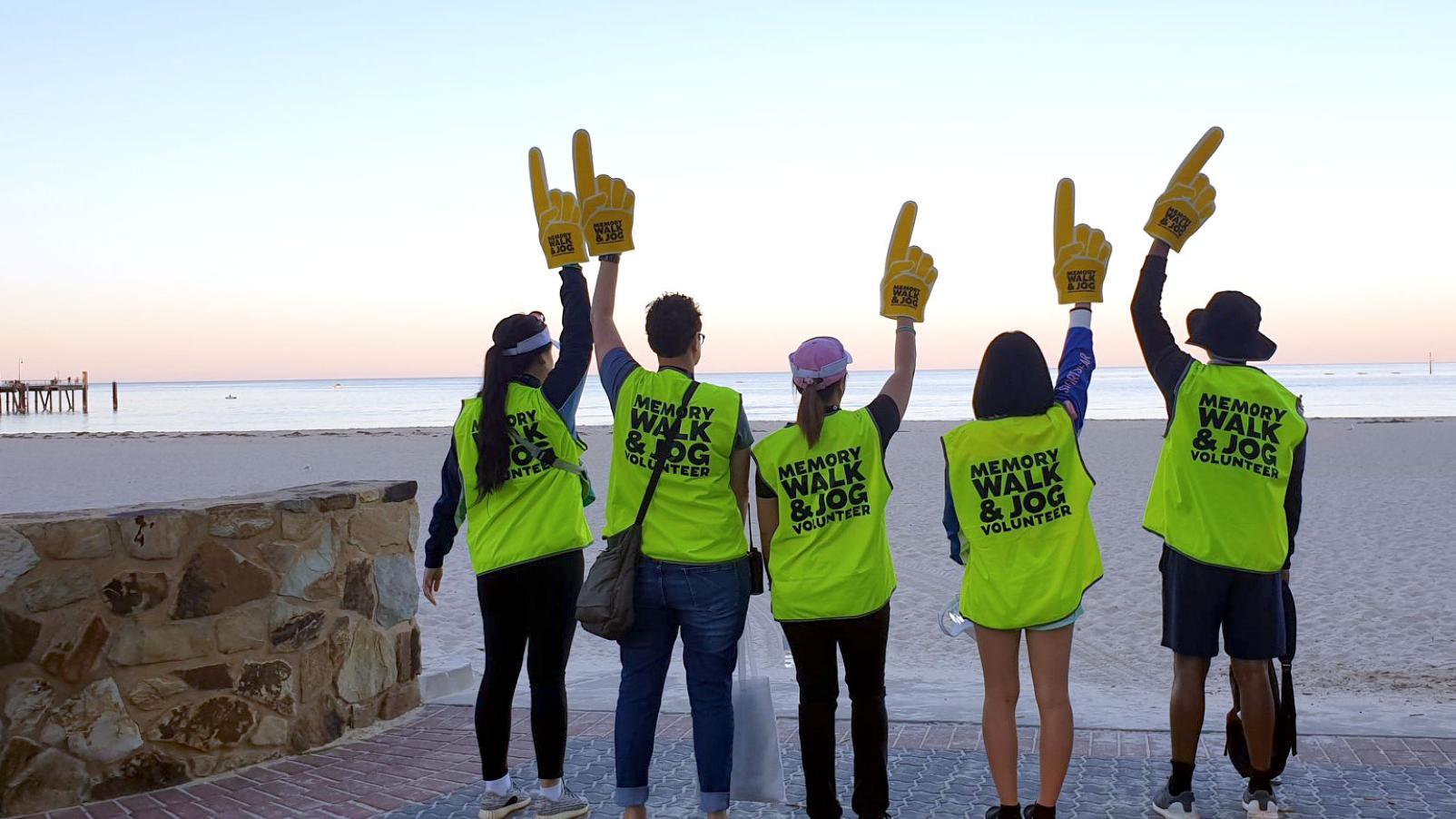 Four people with high visibility yellow vests facing away from the camera, on a beach with large foam fingers raised above their heads