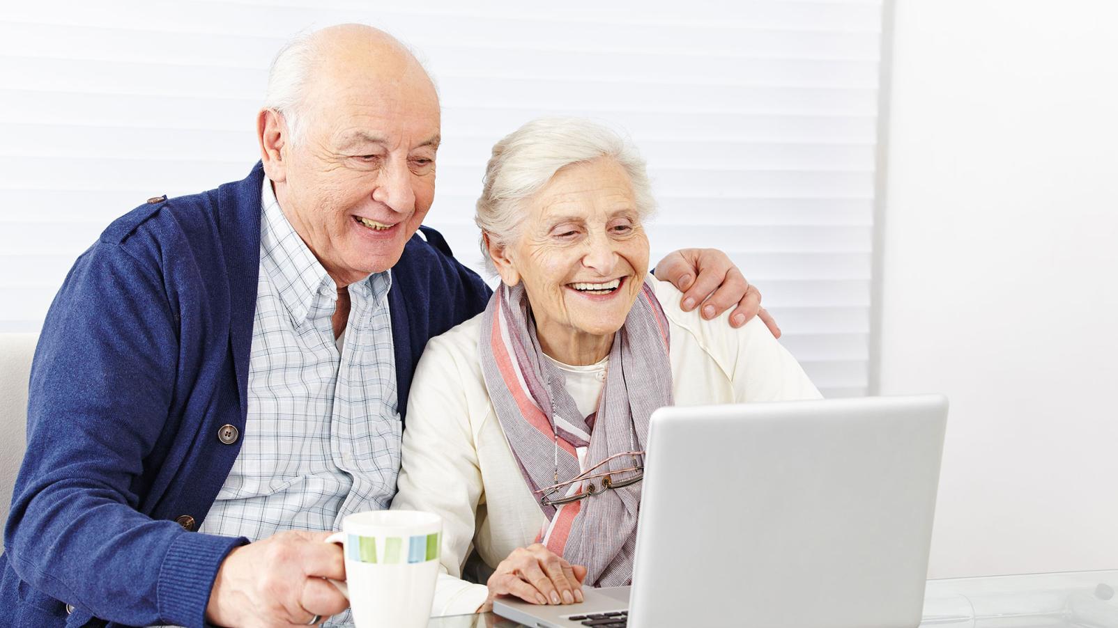 A couple smiling and looking at a laptop on their table. Man has his arm around her.