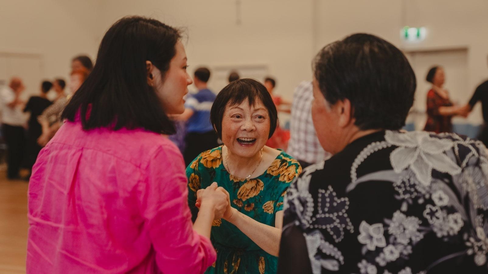 A woman stands smiling in a hall. Two other people hold her hands and talk to her as others gather behind.