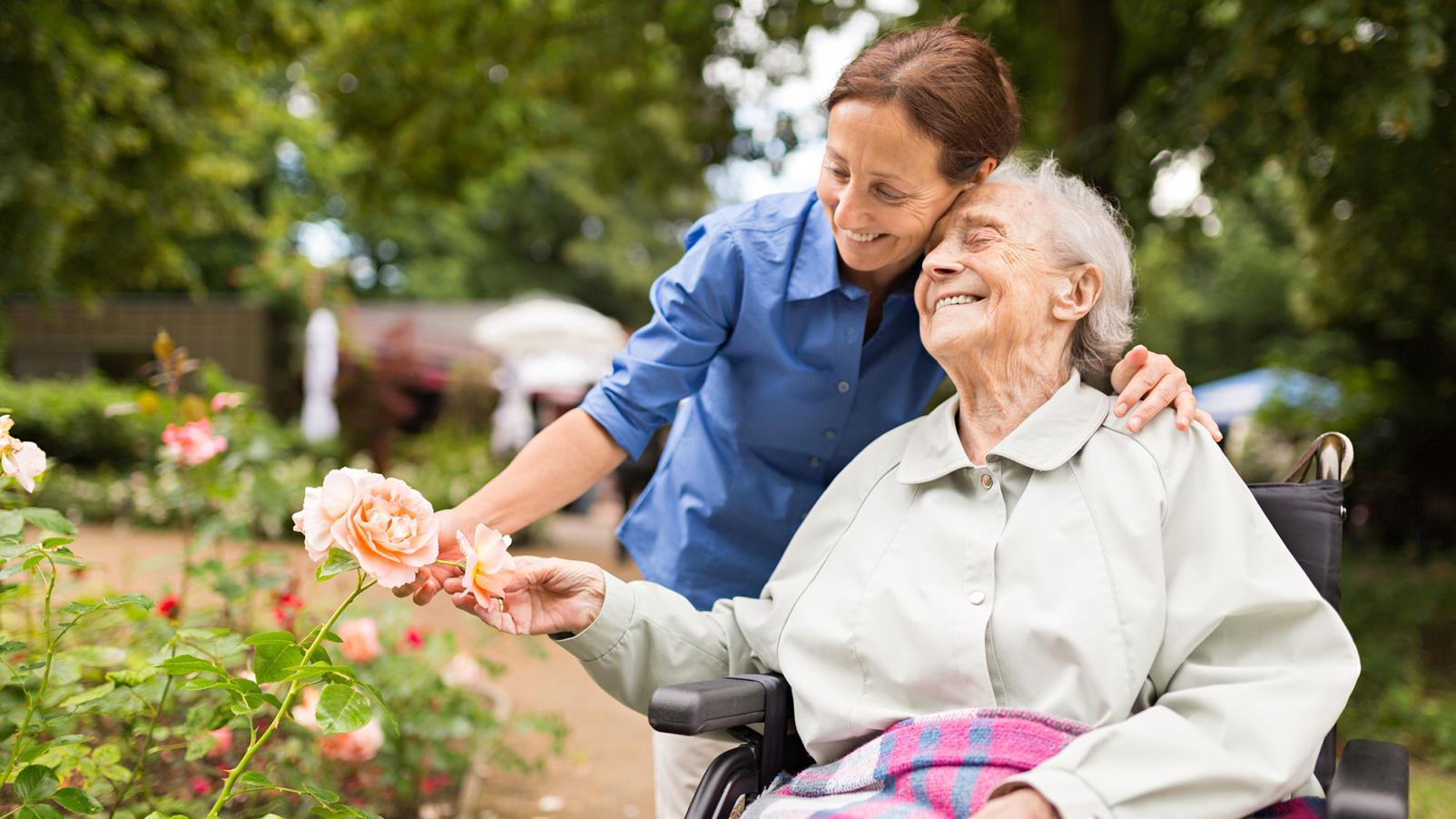 An elderly female in a wheelchair being hugged by a younger female in a garden, both smiling