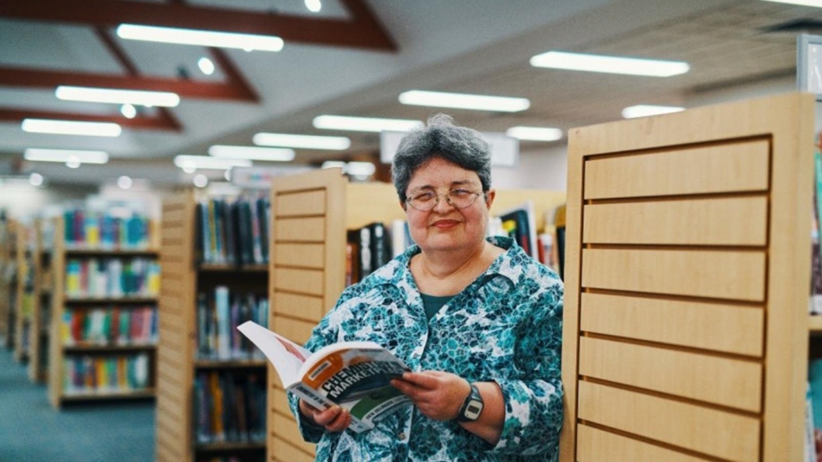 A woman standing in a library holding a book and smiling at the camera