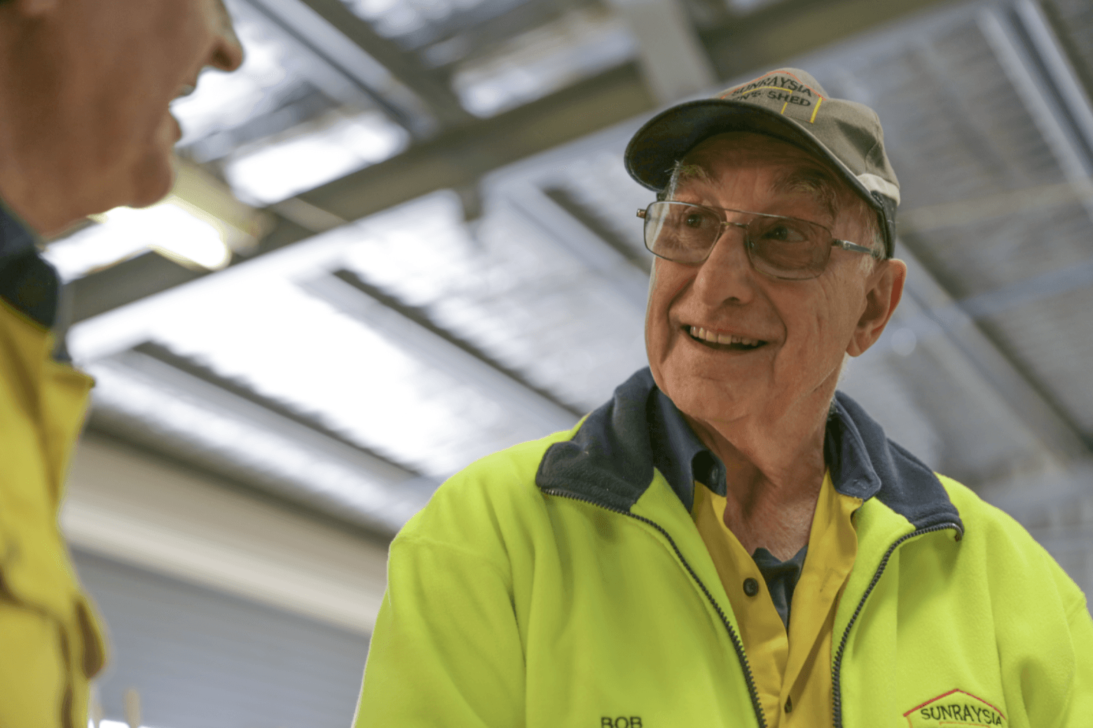 A man called Bob wearing glasses, a hat, and high visibility yellow safety clothing, talking to another man