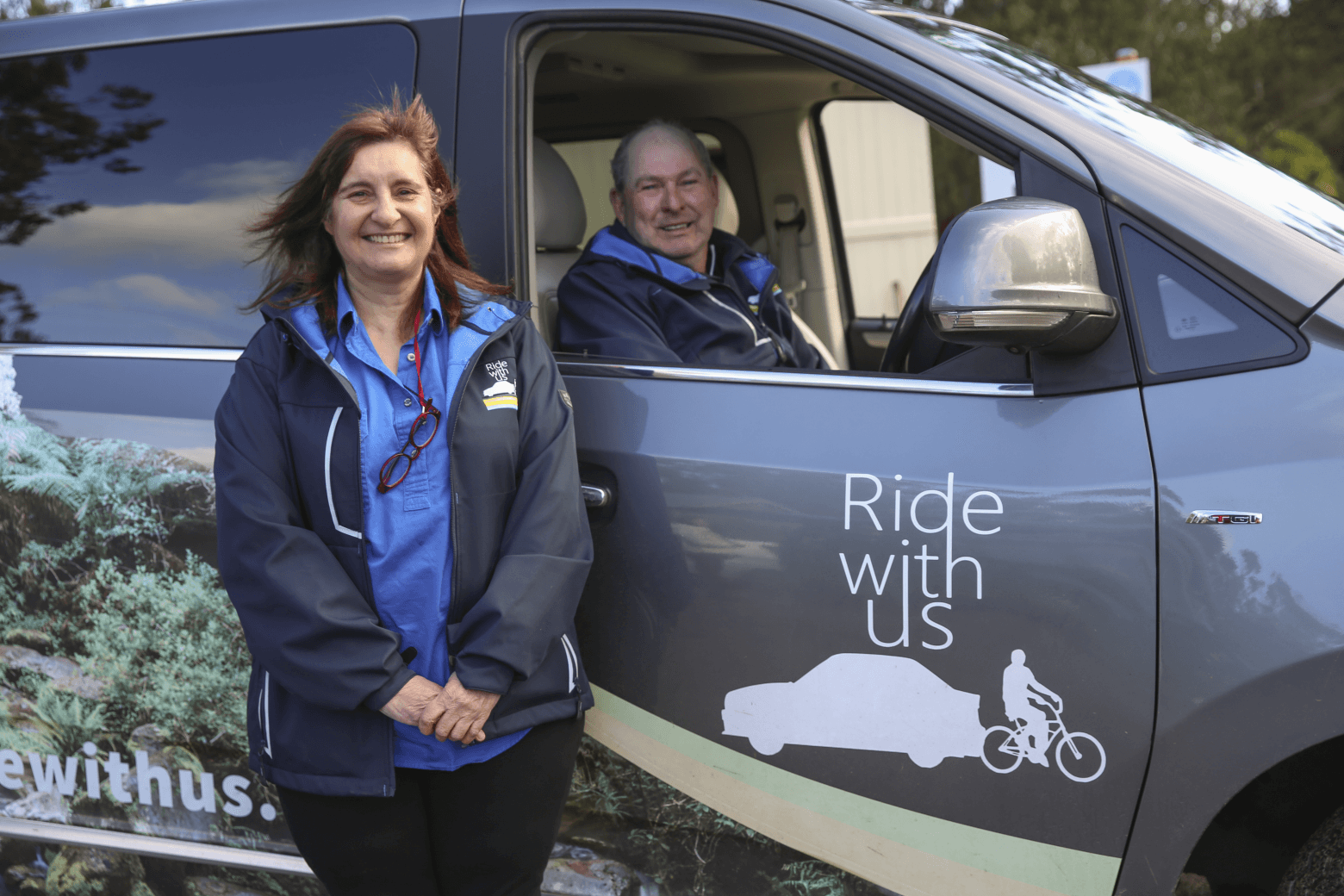 A man sitting in the drivers seat of a car with the logo saying ride with us. Logo has a car and a bike. Sandra is standing in front of the car.