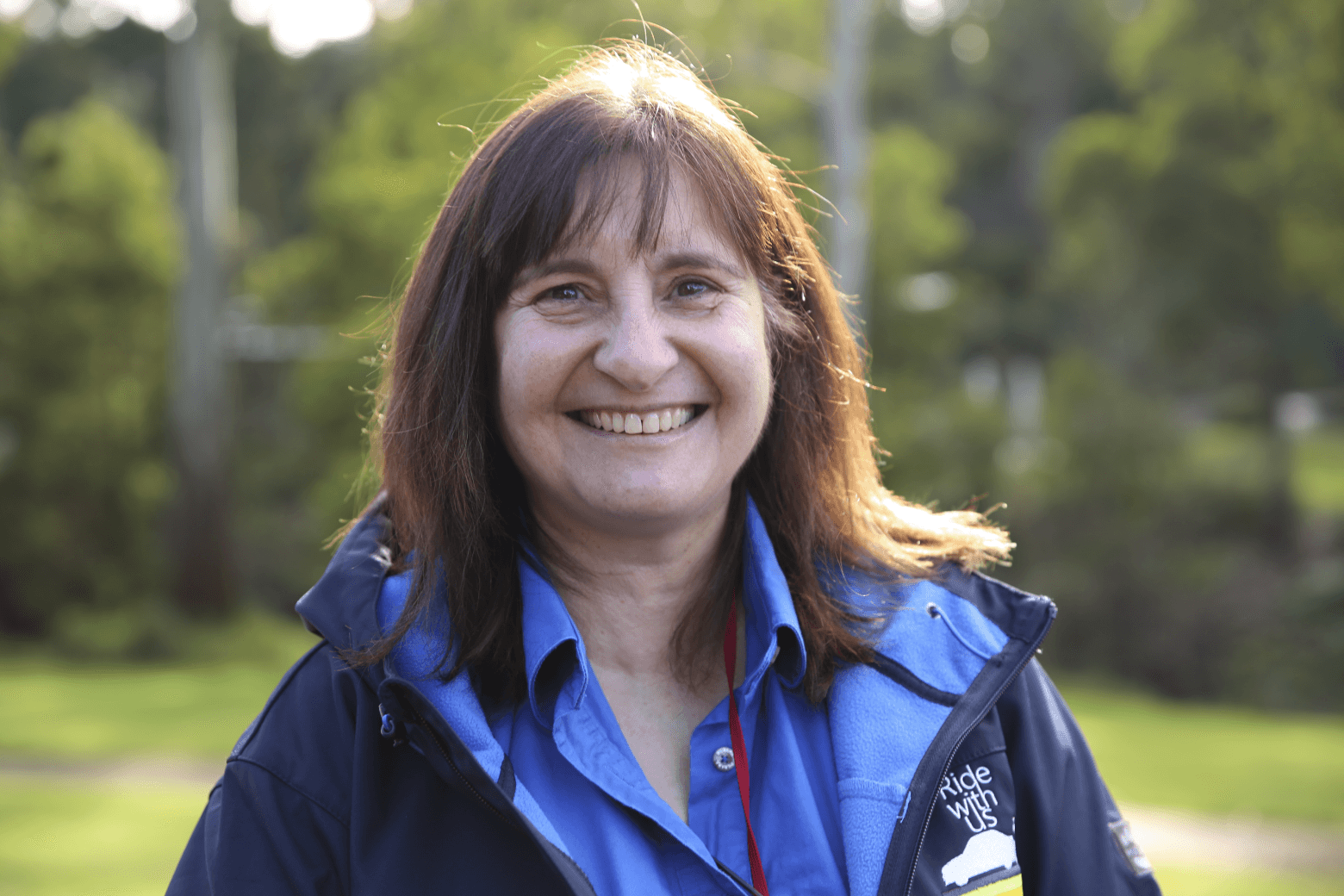 A woman standing outside in the bush smiling at the camera wearing a jacket with a logo that says ride with us