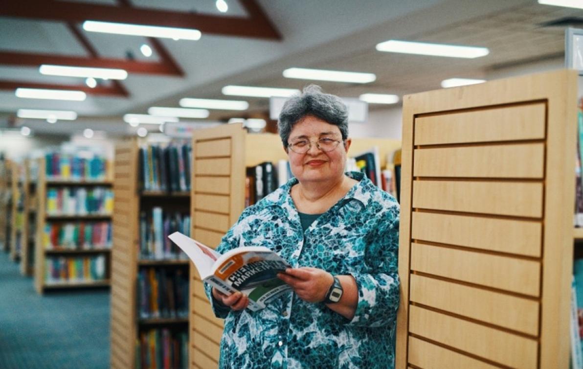 A woman standing in a library holding a book and smiling at the camera