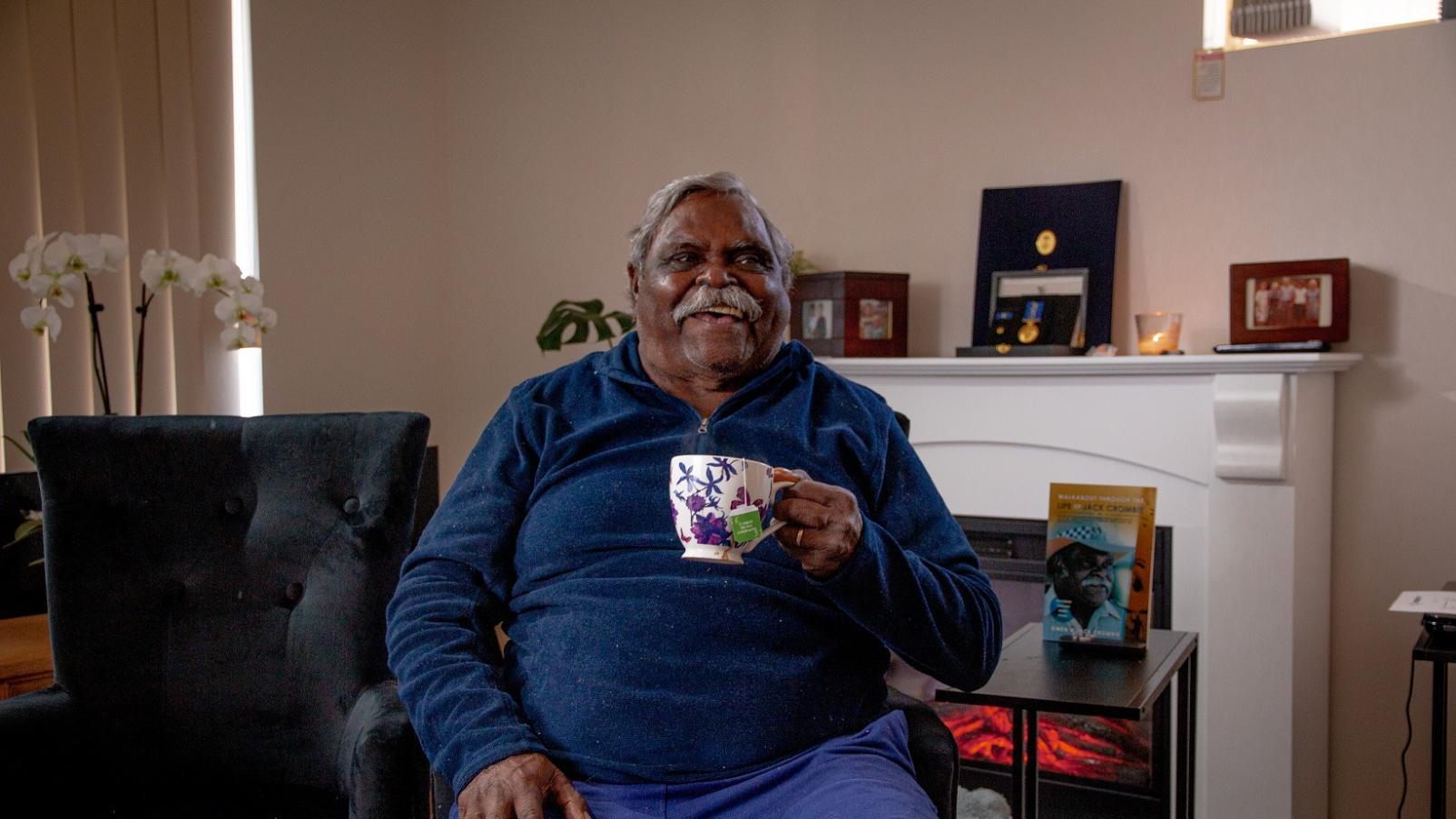A man sitting in his living room holidng a coffee mug and smiling