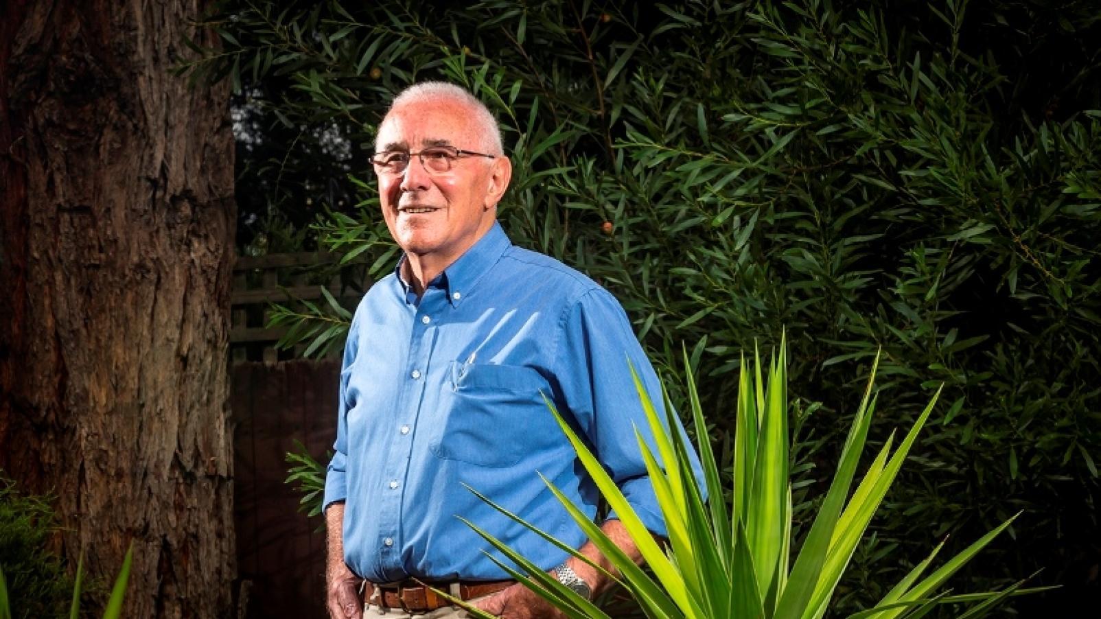 A man wearing a blue shirt standing in his garden with hands in pockets and smiling