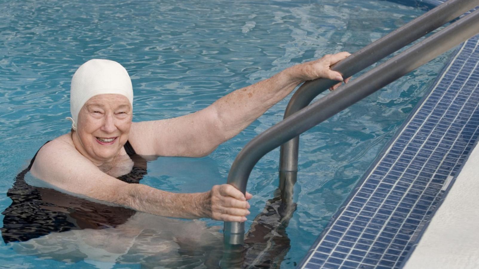 A women in swimwear including a swimmers cap climbing out of a swimming pool and smiling at the camera