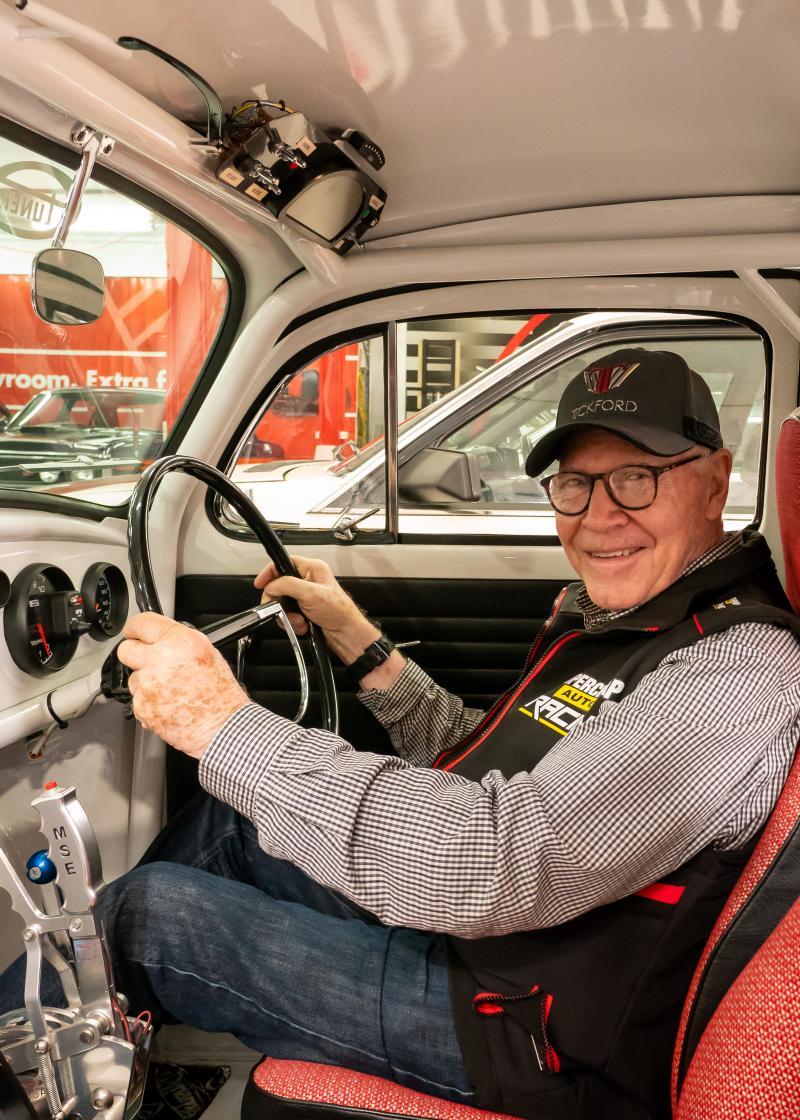 Allan Moffat sitting in the driver's seat of an automobile with his hands on the steering wheel and smiling at the camera.