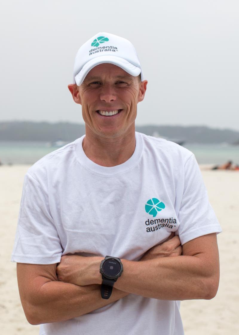 Andrew 'Reidy' Reid, looking at the camera, smiling with his arms folded, wearing a white Dementia Australia hat and a white Dementia Australia t-shirt.