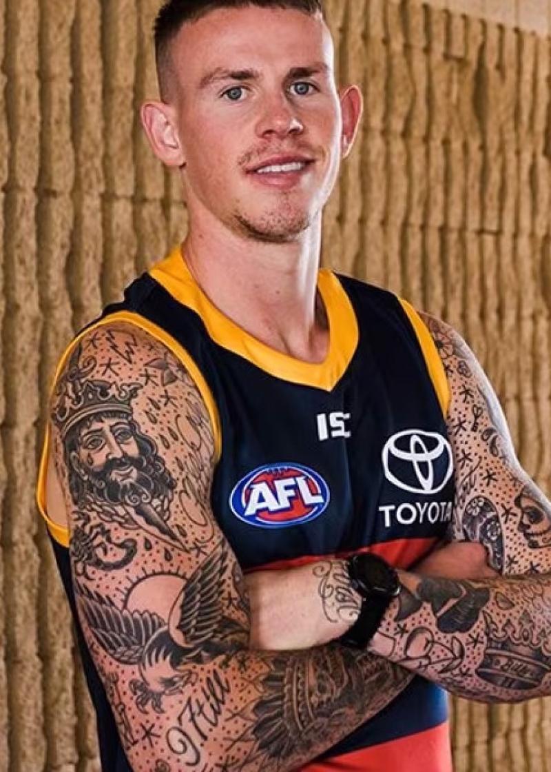 Ben Crocker looking at the camera with his arms folded, smiling, and wearing an Adelaide Crows football jumper.