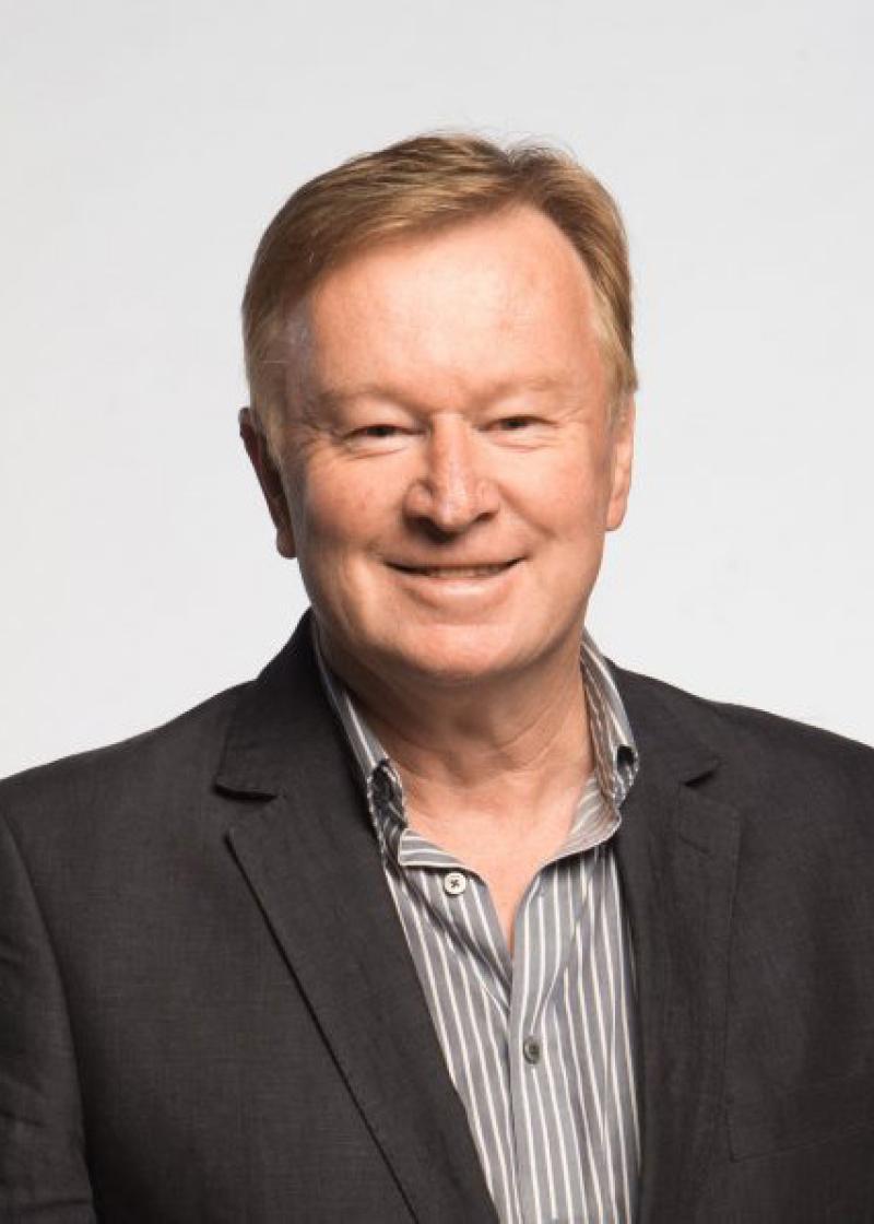 Denis Walter OAM looking at the camera, smiling, wearing a black suit jacket and a grey and white striped shirt.