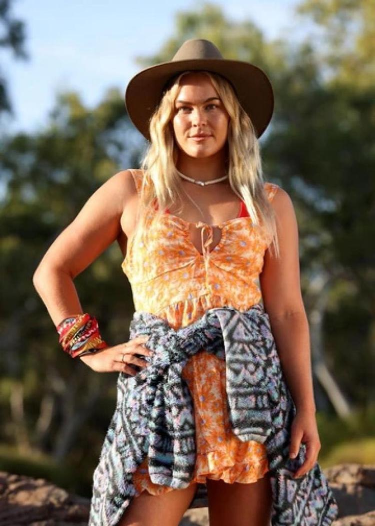 Felicity Palmateer looking at the camera, wearing an akubra hat, an orange floral print dress and a grey and white jumper tied around her waist.