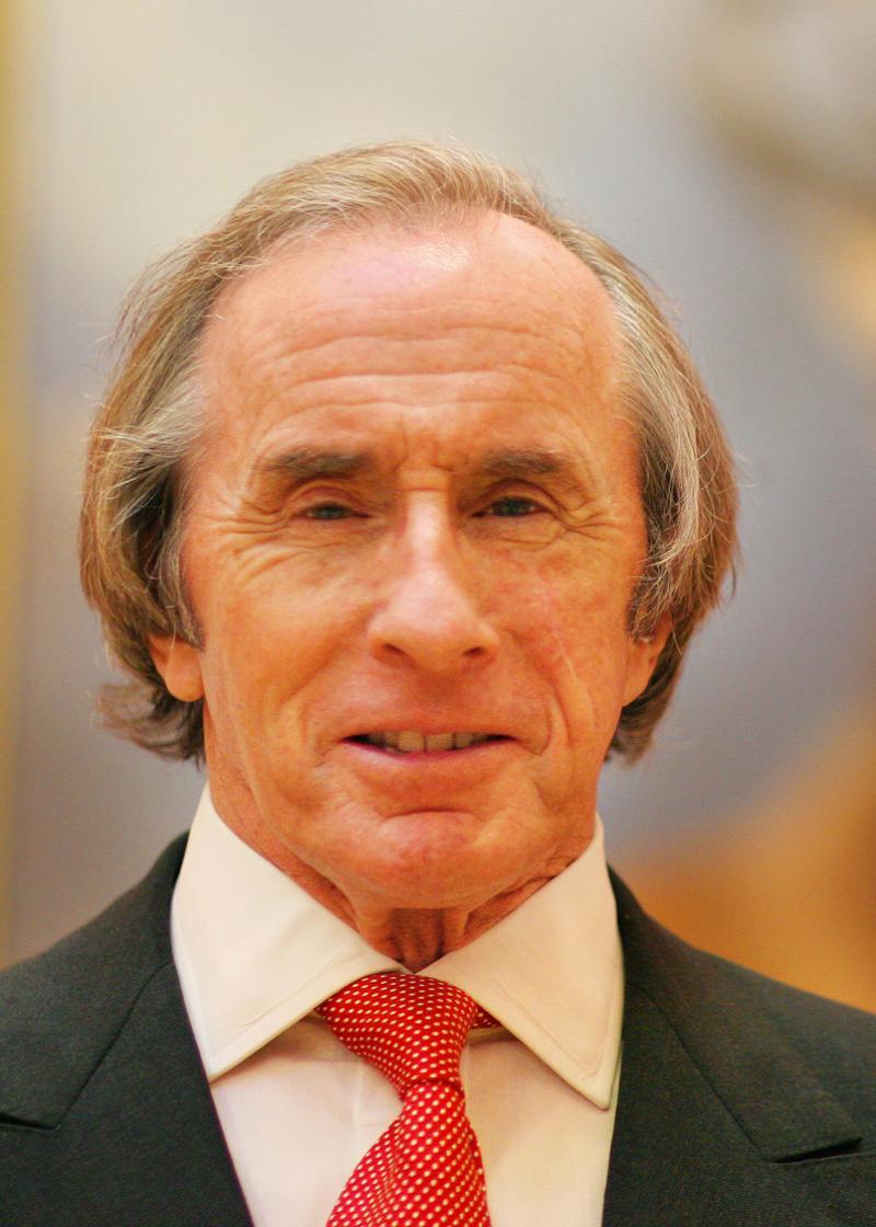 Sir Jackie Stewart OBE looking at the camera wearing a grey suit, white shirt and red tie.