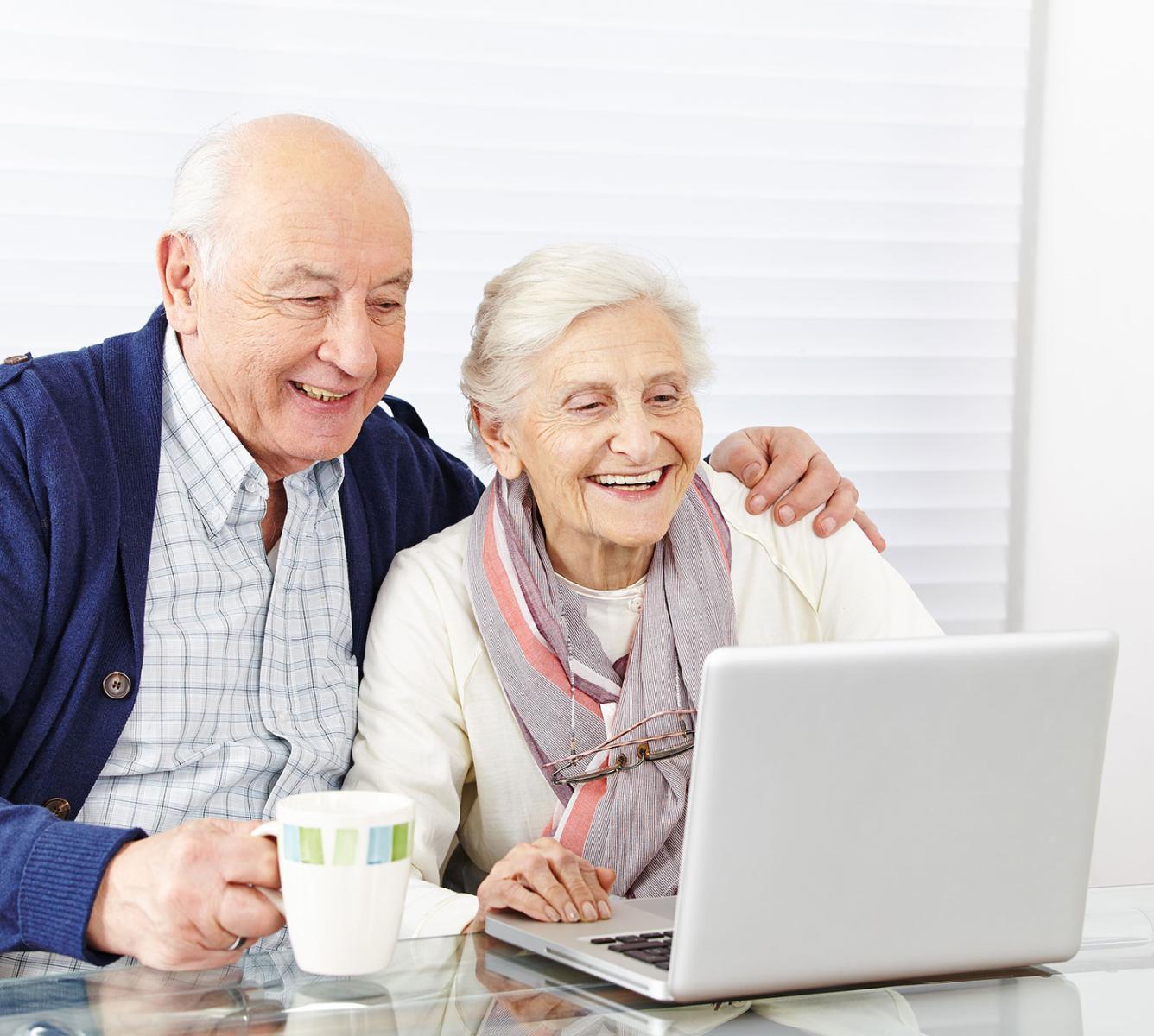 A couple smiling and looking at a laptop on their table. Man has his arm around her.