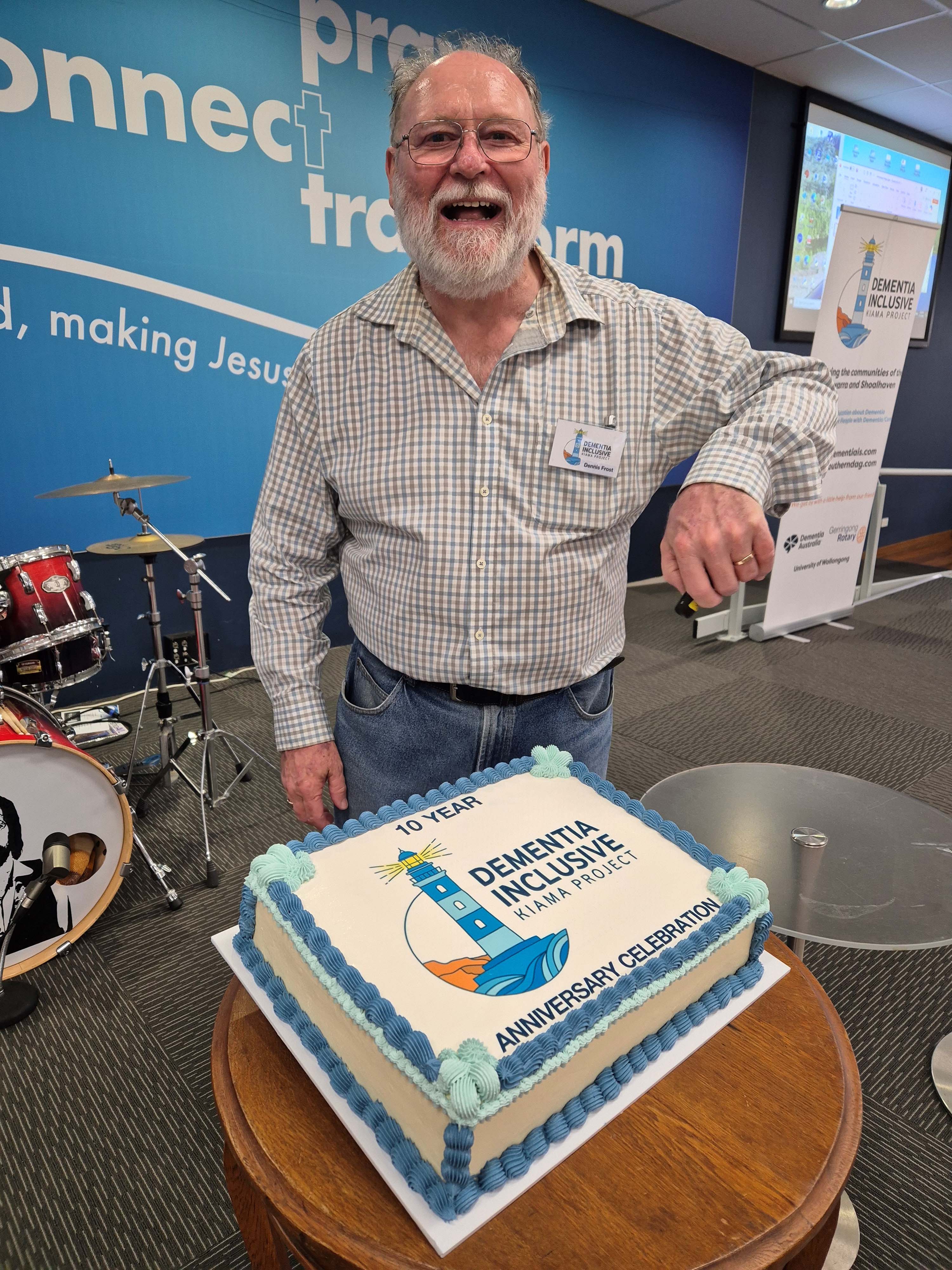 Dementia Advocate Dennis Frost at the Kiama 10 year anniversary celebration, looking at the camera and smiling while standing in front of a cake.